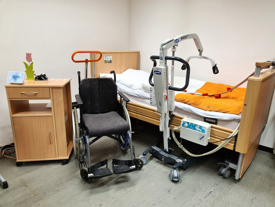 Ein Raum mit einem Pflegebett, einem Rollstuhl und weiteren Hilfen.