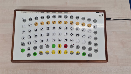 Tastatur mit einer harten Aluminiumplatte und einem Echtholzrahmen. Die Tasten sind vergrößert und liegen tiefer. Sie sind mit weitem Abstand voneinander in der Form eines Halbkreises angeordnet. Einige Tasten sind farbig.
