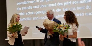 Frau Dr. Susanne Dirks, Herr Prof. Dr.-Ing. Christian Bühler und   Frau Gülistan Demirbilek stehen auf einer Bühne. Sie halten jeweils einen Blumenstrauß in der Hand. Sie lächeln sich an.