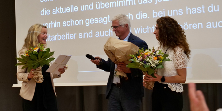 Frau Dr. Susanne Dirks, Herr Prof. Dr.-Ing. Christian Bühler und   Frau Gülistan Demirbilek stehen auf einer Bühne. Sie halten jeweils einen Blumenstrauß in der Hand. Sie lächeln sich an.