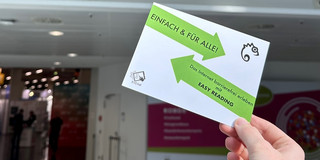 Eine Hand hält einen Flyer, auf dem es um Easy Reading geht. Im Hintergrund ist ein Banner der Werkstätten:messe zu sehen, das herzlich willkommen heißt.