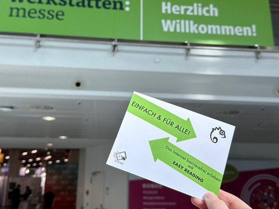 Eine Hand hält einen Flyer, auf dem es um Easy Reading geht. Im Hintergrund ist ein Banner der Werkstätten:messe zu sehen, das herzlich willkommen heißt.