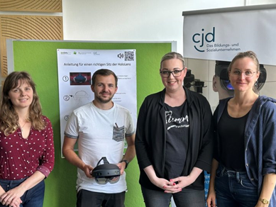 Auf dem Bild sind vier Personen zu sehen, die in einem Raum stehen. Sie lächeln und posieren gemeinsam vor einer grünen Wand, die mit Informationen über die Nutzung von Virtual-Reality-Technologie versehen ist. Eine der Personen hält eine VR-Brille in den Händen. Im Hintergrund ist ein Banner des CJD (Christliches Jugenddorfwerk Deutschlands) sichtbar, das auf Bildungs- und Sozialangebote hinweist. Die Atmosphäre wirkt freundlich und kooperativ, was auf eine Veranstaltung oder einen Workshop hindeutet, der sich mit innovativen Technologien beschäftigt.