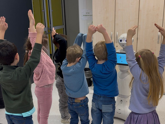 Kinder stehen vor einem humanoiden Roboter und machen gemeinsam Armbewegungen nach, vermutlich während eines Tanz- oder Bewegungsangebots.