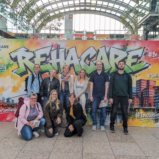 Neun Personen, teilweise stehend und kniend, befinden sich vor dem Eingangsplakat der Messe Rehacare.