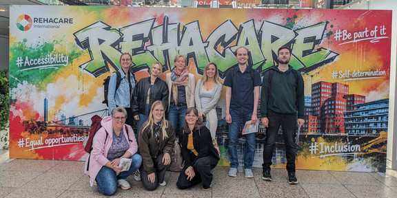 Neun Personen, teilweise stehend und kniend, befinden sich vor dem Eingangsplakat der Messe Rehacare.