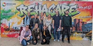 Neun Personen, teilweise stehend und kniend, befinden sich vor dem Eingangsplakat der Messe Rehacare.
