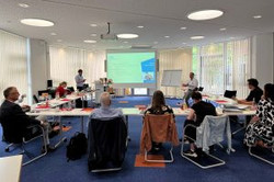 mehrere Personen sitzen in U-Form an Tischen und schauen auf eine Leinwand. Vorne neben der Leinwand stehen zwei Personen