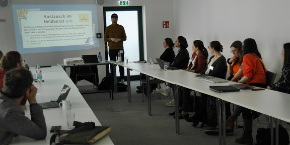 In einem rechteckigen, weiß gestrichenen Konferenzraum findet ein Workshop statt. Etwa zwölf Personen sitzen an drei Seiten eines U-förmig aufgestellten Tisches und blicken auf eine Präsentation, die an die Wand projiziert wird. Die präsentierende Person steht an der offenen Seite des Tisches, nahe der Wand mit der Projektion, trägt einen braunen Pullover und eine dunkle Baseballkappe. Auf der Wand ist eine Folie mit dem Titel „Austausch im Heldenrat (3/3)“ zu sehen. Darunter stehen zwei Fragestellungen zum Einsatz von Serious Play und anderen Methoden in partizipativen Projekten. Die Teilnehmenden sind gemischt, wirken konzentriert und hören aufmerksam zu. Einige haben Laptops, Notizblöcke, Tablets oder Wasserflaschen vor sich. In der Mitte des Tisches steht ein Beamer. Die Atmosphäre ist ruhig und professionell.
