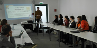 In einem rechteckigen, weiß gestrichenen Konferenzraum findet ein Workshop statt. Etwa zwölf Personen sitzen an drei Seiten eines U-förmig aufgestellten Tisches und blicken auf eine Präsentation, die an die Wand projiziert wird. Die präsentierende Person steht an der offenen Seite des Tisches, nahe der Wand mit der Projektion, trägt einen braunen Pullover und eine dunkle Baseballkappe. Auf der Wand ist eine Folie mit dem Titel „Austausch im Heldenrat (3/3)“ zu sehen. Darunter stehen zwei Fragestellungen zum Einsatz von Serious Play und anderen Methoden in partizipativen Projekten. Die Teilnehmenden sind gemischt, wirken konzentriert und hören aufmerksam zu. Einige haben Laptops, Notizblöcke, Tablets oder Wasserflaschen vor sich. In der Mitte des Tisches steht ein Beamer. Die Atmosphäre ist ruhig und professionell.
