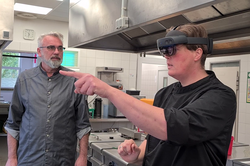 Zwei Personen stehen in einer Küche. Eine Person trägt eine HoloLens und streckt den Arm und Zeigefinger nach vorne aus