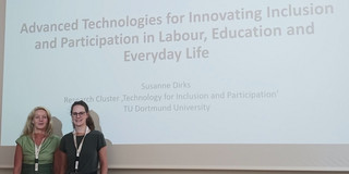 Das Bild zeigt zwei Frauen ( Susanne Dirks links und zu ihrer rechten Leevke Wilkens), die auf einer Konferenz vor einer Präsentationsfolie stehen. Die Präsentationsfolie gehört zur ICCHP (International Conference on Computers Helping People with Special Needs) und hat den Titel "Advanced Technologies for Innovating Inclusion and Participation in Labour, Education and Everyday Life". Unter dem Titel steht der Name der Vortragenden, Susanne Dirks, die am Forschungscluster "Technology for Inclusion and Participation" der TU Dortmund University tätig ist. Beide Frauen tragen Namensschilder, und im Vordergrund rechts befindet sich ein kleiner Tisch mit einem Blumenstrauß.