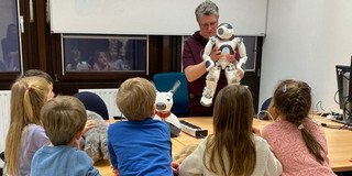 Mehrere Kinder sitzen an einem Tisch und schauen zu, wie eine Person einen kleinen humanoiden Roboter präsentiert und erklärt.
