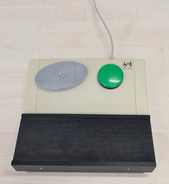 Maus mit zwei Tastern, bestehend aus einer ovalen Taumelplatte links und einem grünen Knopf rechts. Es gibt eine Auflage für den Fuß.