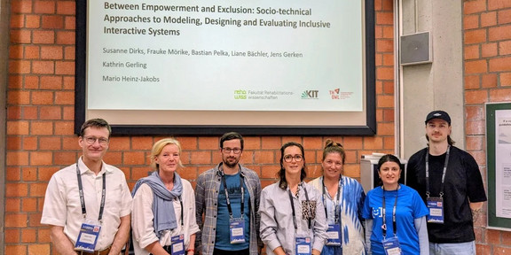 Sieben Personen stehen für ein Gruppenfoto vor einer Projektionsleinwand auf der Konferenz „Mensch und Computer 2026“. Die Leinwand zeigt den Titel eines Workshops zu Empowerment, Exklusion und der Gestaltung inklusiver interaktiver Systeme.