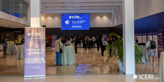  Menschen befinden sich in einem großen Konferenzsaal mit hohen weißen Säulen und einer modernen, offenen Atmosphäre. Im Vordergrund stehen Stehtische mit beigefarbenen Tischdecken, während im Hintergrund ein großer Bildschirm mit dem Text „ICERI 2024“ sichtbar ist. Aufsteller mit dem Schriftzug „WELCOME TO #ICERI2024“ sind ebenfalls prominent platziert. Pflanzen und Glaswände an den Seiten ergänzen die Umgebung