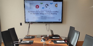 Konferenztisch mit 6 Stühlen. Vor Kopf ist ein Bildschirm an die Wand montiert, auf der eine Easy Reading Präsentation abgebildet ist.