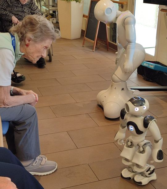 Eine ältere Frau beugt sich nach vorne und betrachtet den kleinen humanoiden Roboter Nao. Hinter ihm steht der größere Roboter Pepper mit gesenktem Kopf.