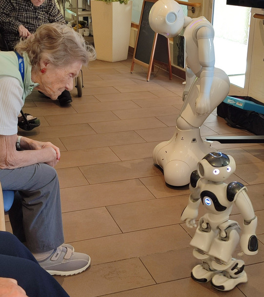 Eine ältere Frau beugt sich nach vorne und betrachtet den kleinen humanoiden Roboter Nao. Hinter ihm steht der größere Roboter Pepper mit gesenktem Kopf.
