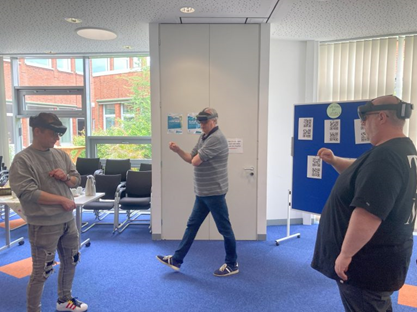 Das Bild zeigt drei Personen, die in einem Raum stehen und Virtual-Reality-Brillen tragen. Eine Person in der Mitte bewegt sich aktiv, während die anderen beiden stehen und beobachten. Der Raum ist hell erleuchtet und hat große Fenster, durch die Tageslicht strömt. Im Hintergrund sind Möbel wie Stühle und Tische sichtbar, die auf eine entspannte Umgebung hindeuten. An einer Wand befindet sich ein Informationsboard mit QR-Codes. Die Szene vermittelt einen Eindruck von Interaktivität und technologischem Lernen.