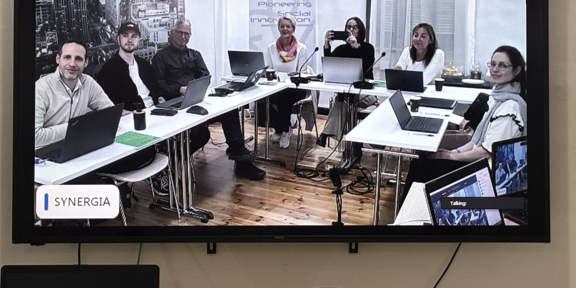 Ein Bildschirm, auf dem die teilnehmenden Personen des UPowerWAD-Projekt-Konsortiums zu sehen sind. Sie sitzen an Tischen und haben ihre Laptops vor sich.