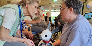 Eine Vorführende hält den Roboter MiRo vor eine ältere Frau und spricht mit ihr. Die Frau beugt sich interessiert nach vorne.
