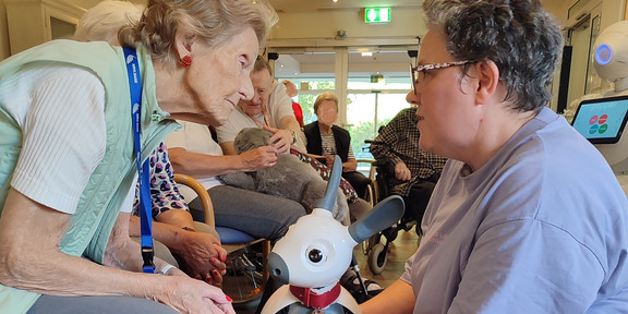 Eine Vorführende hält den Roboter MiRo vor eine ältere Frau und spricht mit ihr. Die Frau beugt sich interessiert nach vorne.