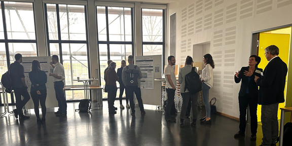 Mehrere Personen stehen in einem hellen Raum mit großen Fenstern, durch die Tageslicht einfällt. Sie betrachten Poster an Stellwänden oder unterhalten sich. Die Atmosphäre wirkt informell und konzentriert. Einige Teilnehmer tragen Rucksäcke, und Tische sind am Rand des Raumes aufgestellt.