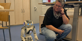 Eine Person hockt in einem Büro auf Augenhöhe mit einem kleinen humanoiden Roboter. Im Hintergrund sind Schreibtische mit Computermonitoren, Schränke, Pflanzen und große Fenster zu sehen.