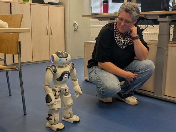 Eine Person hockt in einem Büro auf Augenhöhe mit einem kleinen humanoiden Roboter. Im Hintergrund sind Schreibtische mit Computermonitoren, Schränke, Pflanzen und große Fenster zu sehen.