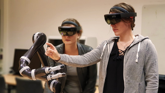Zwei weibliche Personen, die eine HoloLens tragen und vor einem Roboterarm stehen. Die Person rechts zeigt auf den Arm. 