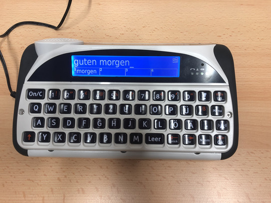Lightwriter Gerät mit Tastatur und Display. Auf der Rückseite ist ein zweites Display und ein Lautsprecher. Das Gerät hat einen Kabelanschluss.