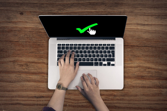 Eine Person tippt mit der linken Hand auf der Tastatur eines Laptops und fährt mit der rechten Hand über das Touchpad. Auf dem Bildschirm ist der Mauszeiger auf einen grünen Haken gerichtet.