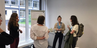 Marie-Christin Lueg steht neben einem Poster. Vier Frauen im Raum schauen sie an.