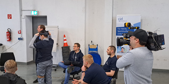 Ein Kameramann filmt eine Gruppe von Personen. Vier Personen sitzen auf Stühlen in einem Halbkreis. Eine Person steht in der Mitte und trägt eine Hololens. 