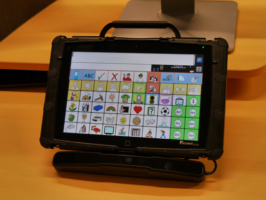 schwarzes Gerät mit Schutzhülle und einem Display mit Touchscreen, auf dem verschiedene Symbole und ein Textfeld sind. Das Gerät hat oben einen Tragegriff und unten eine Halterung.