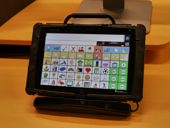 Talker Accent 1000 schwarzes Gerät mit Schutzhülle und einem Display mit Touchscreen, auf dem verschiedene Symbole und ein Textfeld sind. Das Gerät hat oben einen Tragegriff und unten eine Halterung.