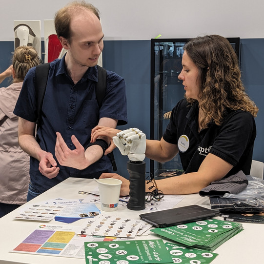 Ein Teammitglied des Fachgebiets Rehabilitationstechnologie erprobt auf der REHACARE die Steuerung einer Handprothese über ein Sensorband am Unterarm. Eine Mitarbeiterin am Messestand unterstützt ihn dabei.