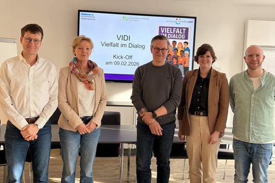 Fünf Personen stehen in einem hellen Besprechungsraum vor einem großen Bildschirm. Auf dem Bildschirm ist die Präsentationsfolie „VIDI – Vielfalt im Dialog“ mit dem Hinweis auf ein Kick-off am 09.02.2026 zu sehen. Außerdem sind die Logos der beteiligten Organisationen sowie eine Illustration mit verschiedenen Personen und dem Schriftzug „Vielfalt im Dialog“ abgebildet. Die Gruppe blickt in die Kamera und wirkt freundlich sowie professionell. Vor ihnen stehen Tische und Stühle eines Seminar- oder Konferenzraums.