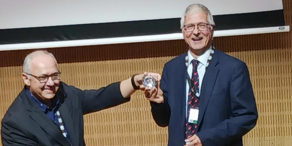 Auf der linken Bildseite ist der amtierende Präsident der AAATE Prof. Georgios Kouroupetroglou zu sehen. Er übergibt den AAATE Diamond Award an Prof. Dr. Christian Bühler, der auf der rechten Bildseite zu sehen ist. Der Award ist ein großes glänzendes Glasobjekt. Porf. Bühler strahlt über das ganze Gesicht. .