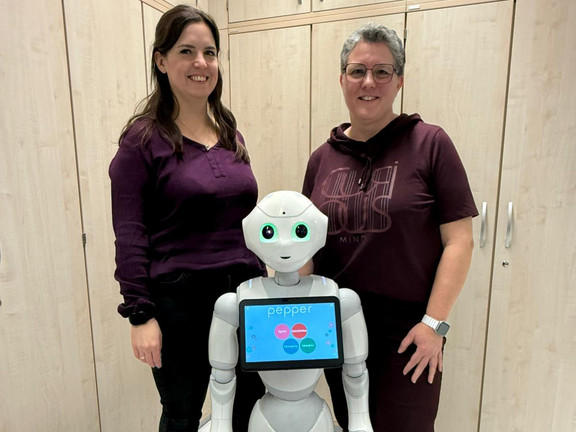 Robo Lab Kita Zwei Erwachsene stehen neben einem humanoiden Roboter und lächeln in die Kamera, im Hintergrund sind Schränke eines Gruppen- oder Technikraums zu sehen.