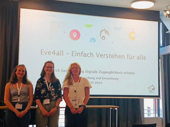 Drei Personen stehen vor einer Leinwand und lächeln in die Kamera. Auf der Leinwand sieht man den Text "Eve4all - Einfach Verstehen für alle".