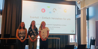 Drei Personen stehen vor einer Leinwand und lächeln in die Kamera. Auf der Leinwand sieht man den Text "Eve4all - Einfach Verstehen für alle".
