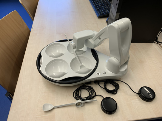 Esshilfe Obi Ein Roboterarm ist an einem Teller mit 4 Schalen befestigt. Zwei Kabel mit jeweils einem Taster sind mit dem Teller verbunden. Es liegt ein Löffel daneben.