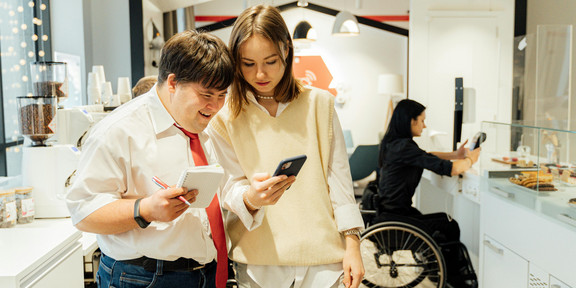 Zwei Personen stehen neben einer Cafétheke und schauen gemeinsam auf ein Smartphone; eine Person mit Notizblock notiert etwas. Im Hintergrund arbeitet eine weitere Person im Rollstuhl an der Theke.
