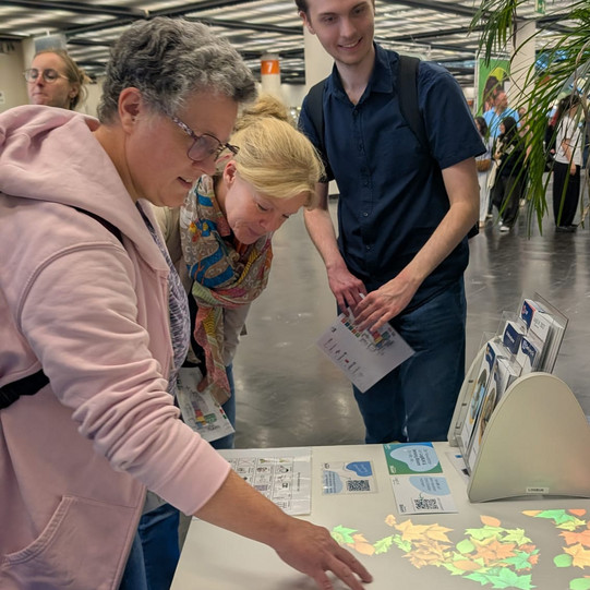 Drei Teammitglieder des Fachgebiets Rehabilitationstechnologie erkunden auf der REHACARE einen interaktiven Projektionstisch. Eine Person bewegt ihre Hand über die auf die Tischfläche projizierten bunten Blätter.