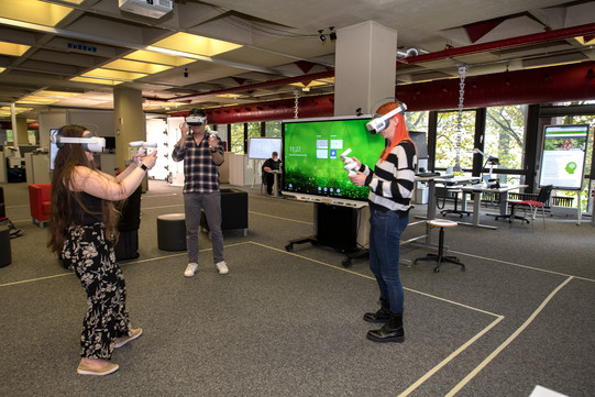 Zwei weibliche Personen und eine männliche Person im Hylec, die VR-Brillen tragen und in den Händen Controller halten. Im Hintergrund sind verschiedene Arbeitsplätze zu sehen.
