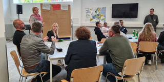  In einem großen hellen Raum sitzen an zwei Tischgruppen jeweils 6 diskutierende Personen unterschiedlichen Alters und Geschlechts. Im Hintergrund ist eine Flip Chart mit Notizen zu sehen. Zwei  Personen, die die Diskussionen leiten, stehen neben den Tischen.