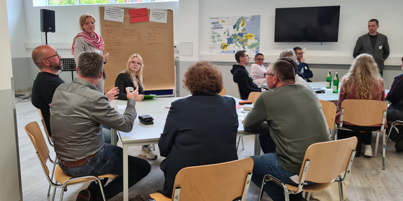  In einem großen hellen Raum sitzen an zwei Tischgruppen jeweils 6 diskutierende Personen unterschiedlichen Alters und Geschlechts. Im Hintergrund ist eine Flip Chart mit Notizen zu sehen. Zwei  Personen, die die Diskussionen leiten, stehen neben den Tischen.