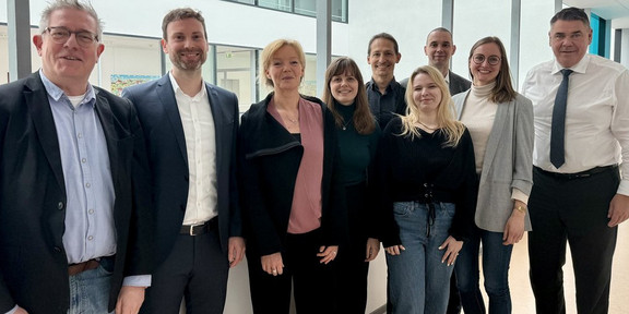 9 Personen verschiedenen Alters und Geschlechts in Smart Causal gekleidet stehen lächelnd nebeneinander auf einem Büroflur.